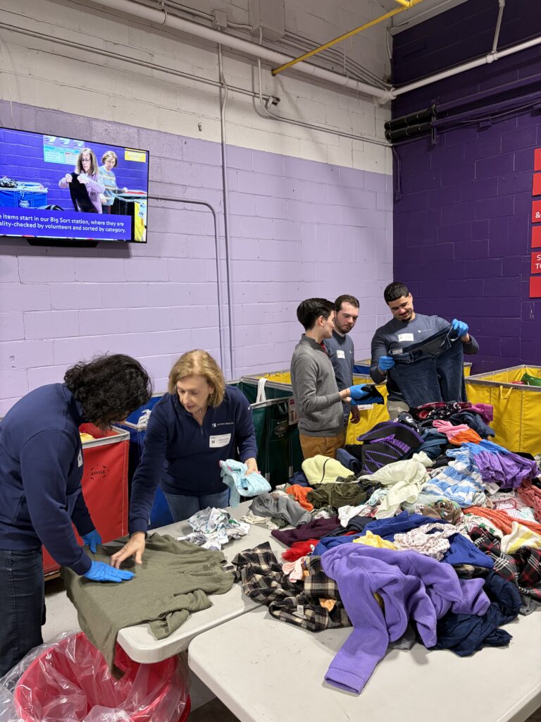 Five people sort donated clothes on tables in a brightly colored room. They wear gloves and appear focused, conveying a sense of teamwork and purpose.