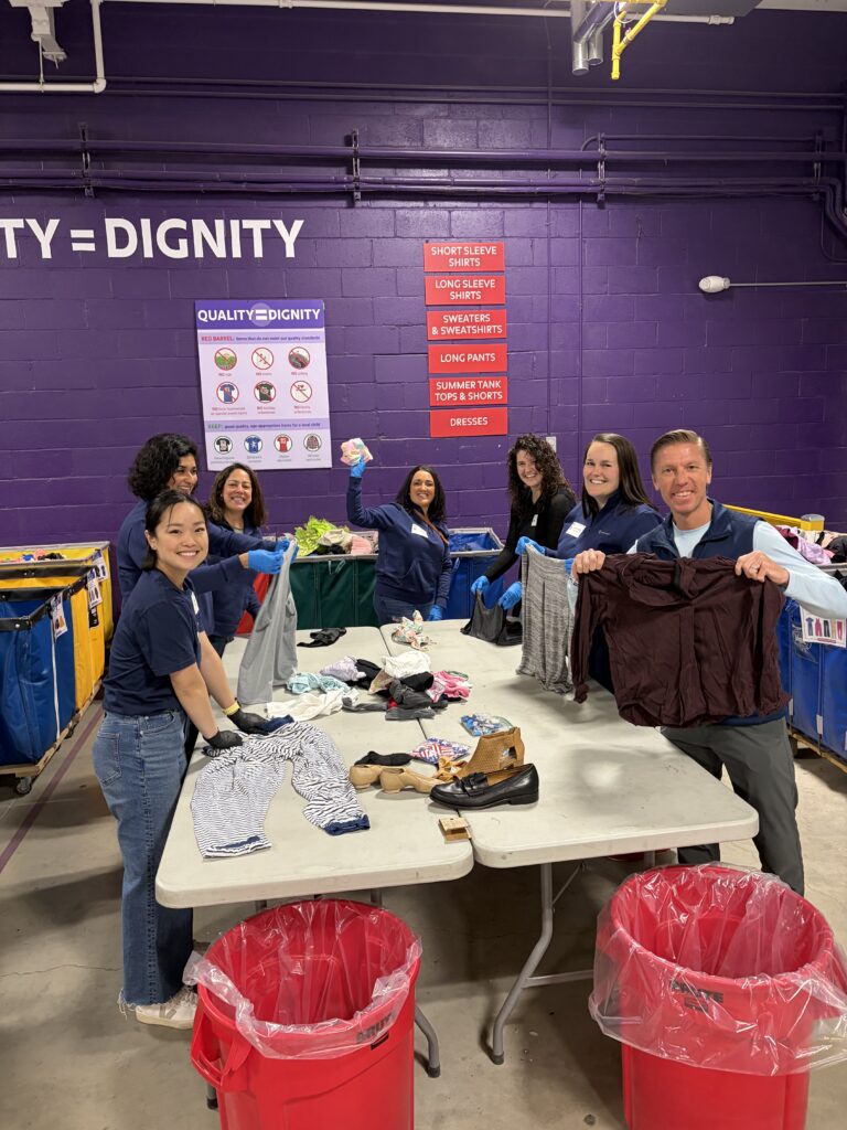 A group of people in navy shirts smiling while sorting clothes at a purple-walled facility. Red bins and posters with sorting guidelines are visible.