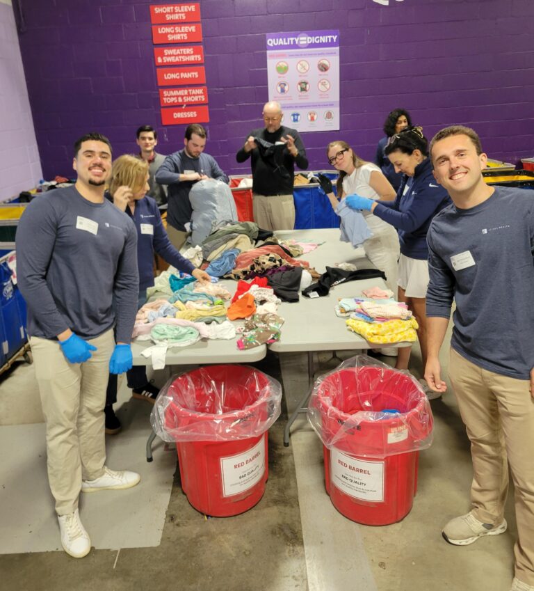 A group of people joyfully sorts clothes on tables in a donation center. Bright red bins are in the foreground, and colorful clothing piles are visible.