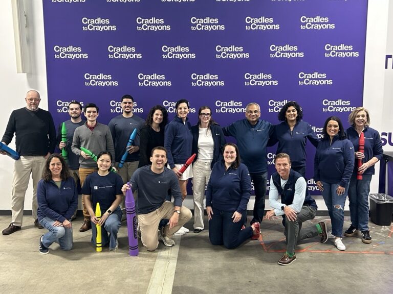 A diverse group of fourteen people, smiling and holding large, colorful crayon props, pose in front of a "Cradles to Crayons" branded backdrop. The atmosphere is cheerful and collaborative.